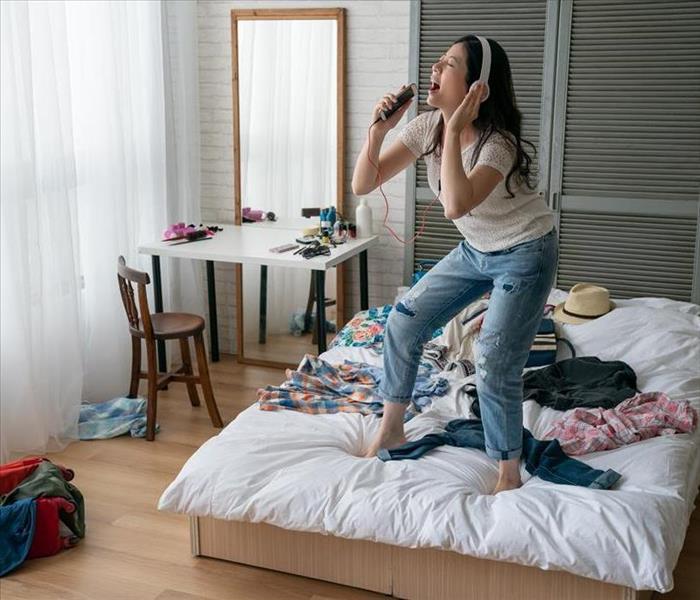 A girl wearing headphones sings into a karaoke microphone while standing on a clothes-covered bed in a messy dorm room